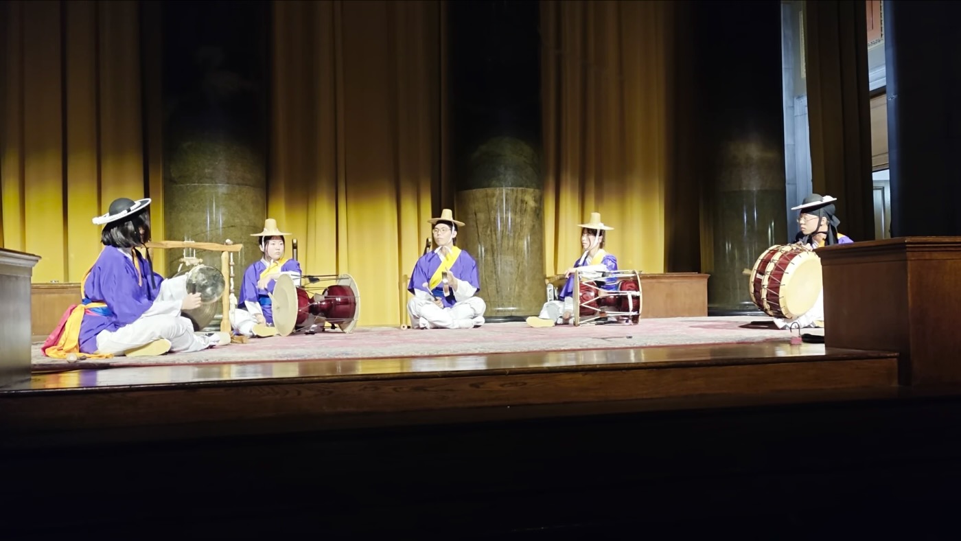 Members in purple performance outfits and hats playing samulnori seated on stage